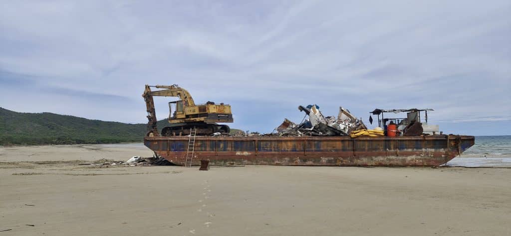 Rusty barge with a large excavator on its deck, piled with debris, on a sandy beach with calm sea and green hills in the background. The vessel appears abandoned and damaged.
