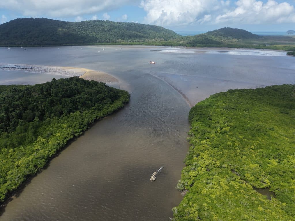 Aerial view of a mangrove estuary with forested banks, calm brown water, and small boats dotting the waterway.