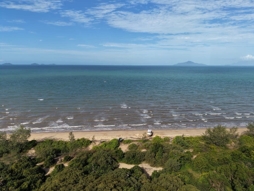 Aerial view of a sandy beach, turquoise sea, and distant islands beneath a blue sky with scattered clouds.
