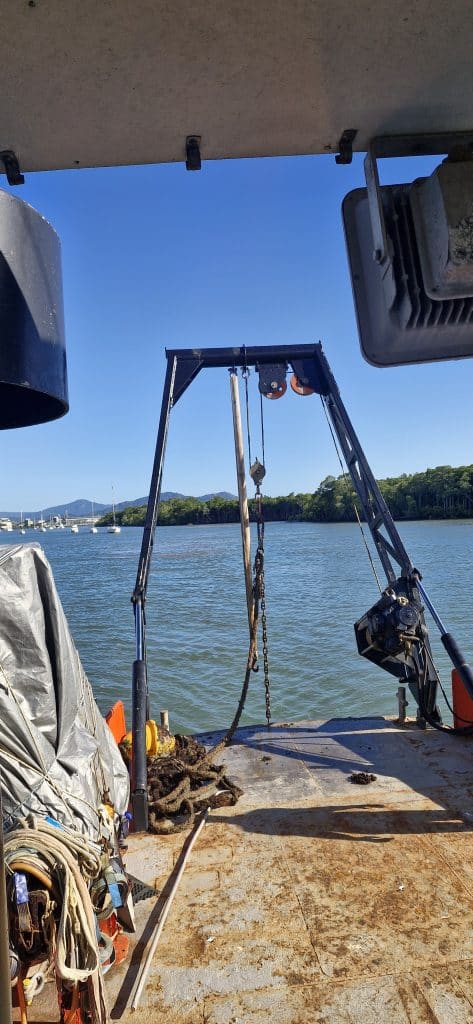 View from a boat deck with a metal hoist frame, rigging, and coiled ropes overlooking calm water and distant shoreline trees.
