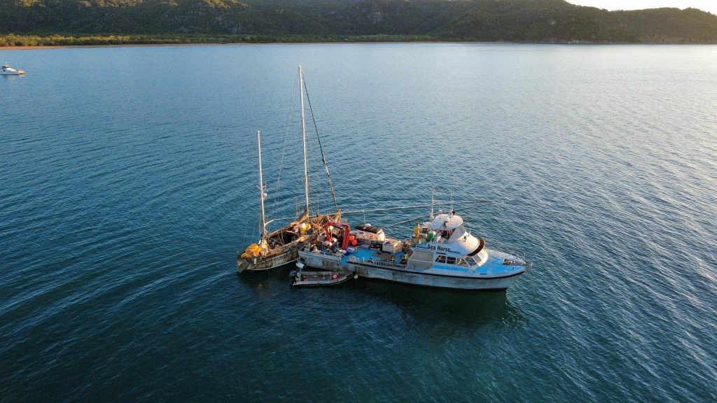Two boats moored together on a calm blue bay with a tree-covered hill in the background.