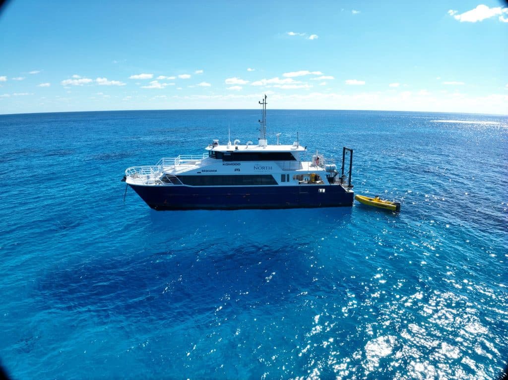 White-and-navy passenger yacht named Komodo on a calm deep-blue sea under a bright sky, with a small yellow dinghy nearby.