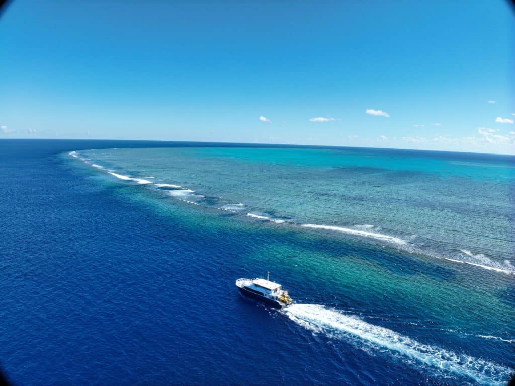 Aerial view of a white yacht cruising in deep blue ocean with white wake, turquoise near-shore water, and clear blue sky above the horizon